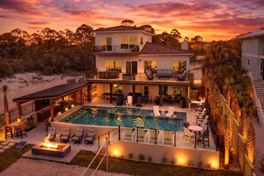 A two-story house with a swimming pool, outdoor seating, and a view of the beach at sunset.