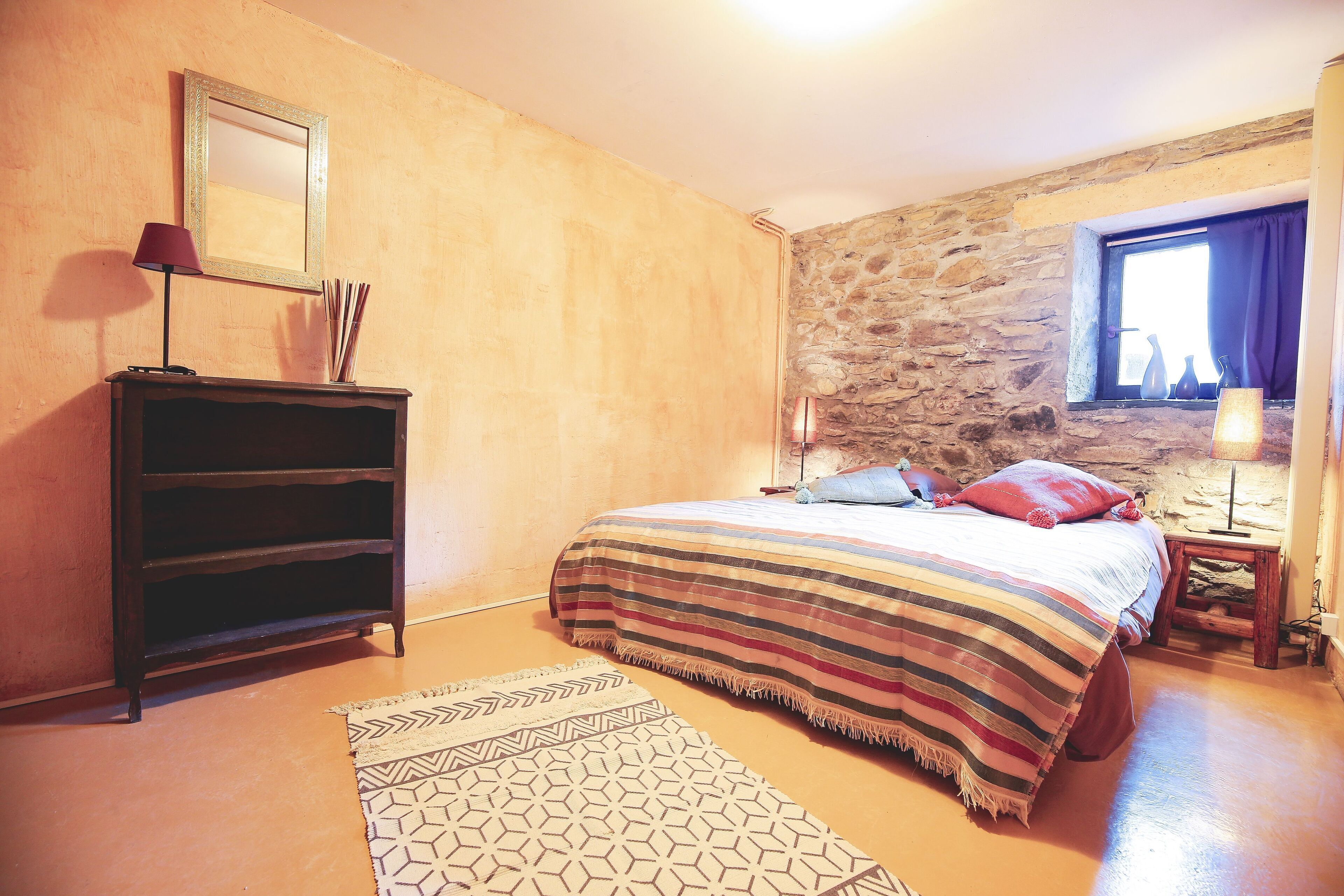 Un dormitorio con cama, cómoda, alfombra, ventana con cortinas y pared de piedra.