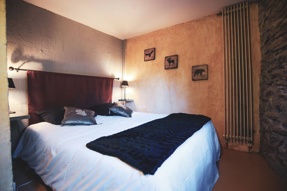 A bedroom with a bed, pillows, a blanket, a nightstand with a lamp, and wall art.