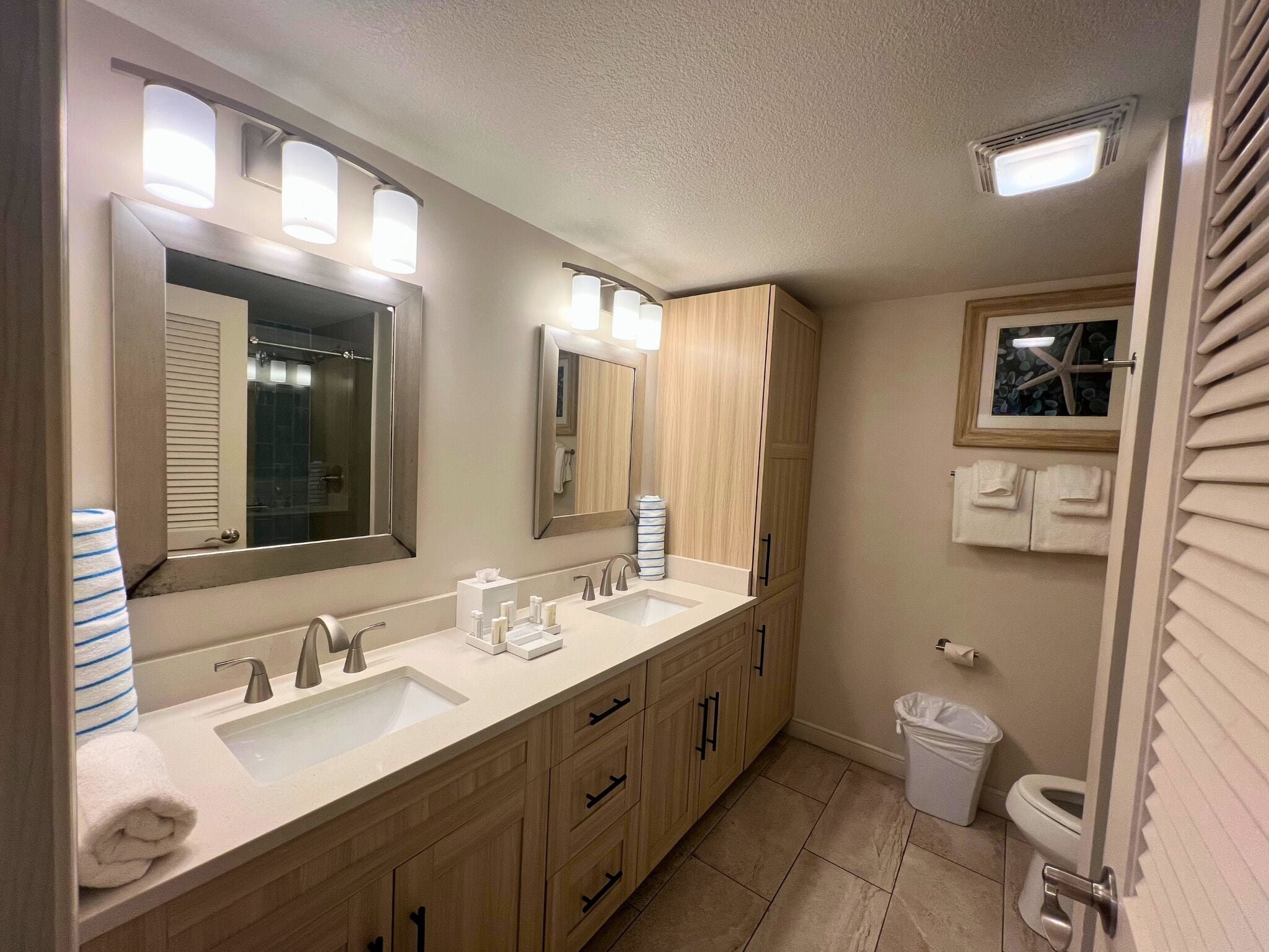 A modern bathroom with a double sink, vanity, and a large mirror. There are wooden cabinets, a trash can, and a toilet visible.