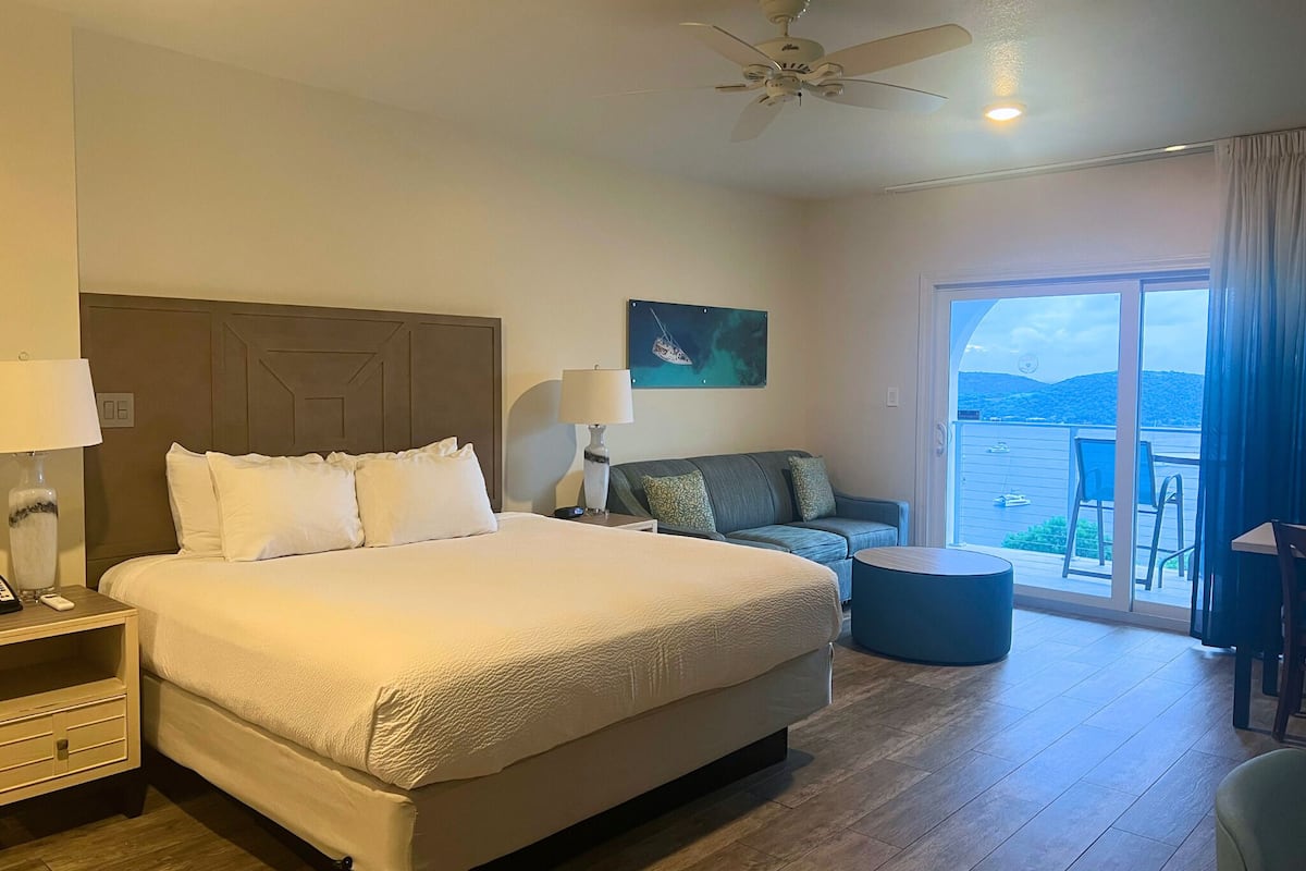 A hotel room with a large bed, a ceiling fan, a painting, a view of the sea, and a balcony with chairs.