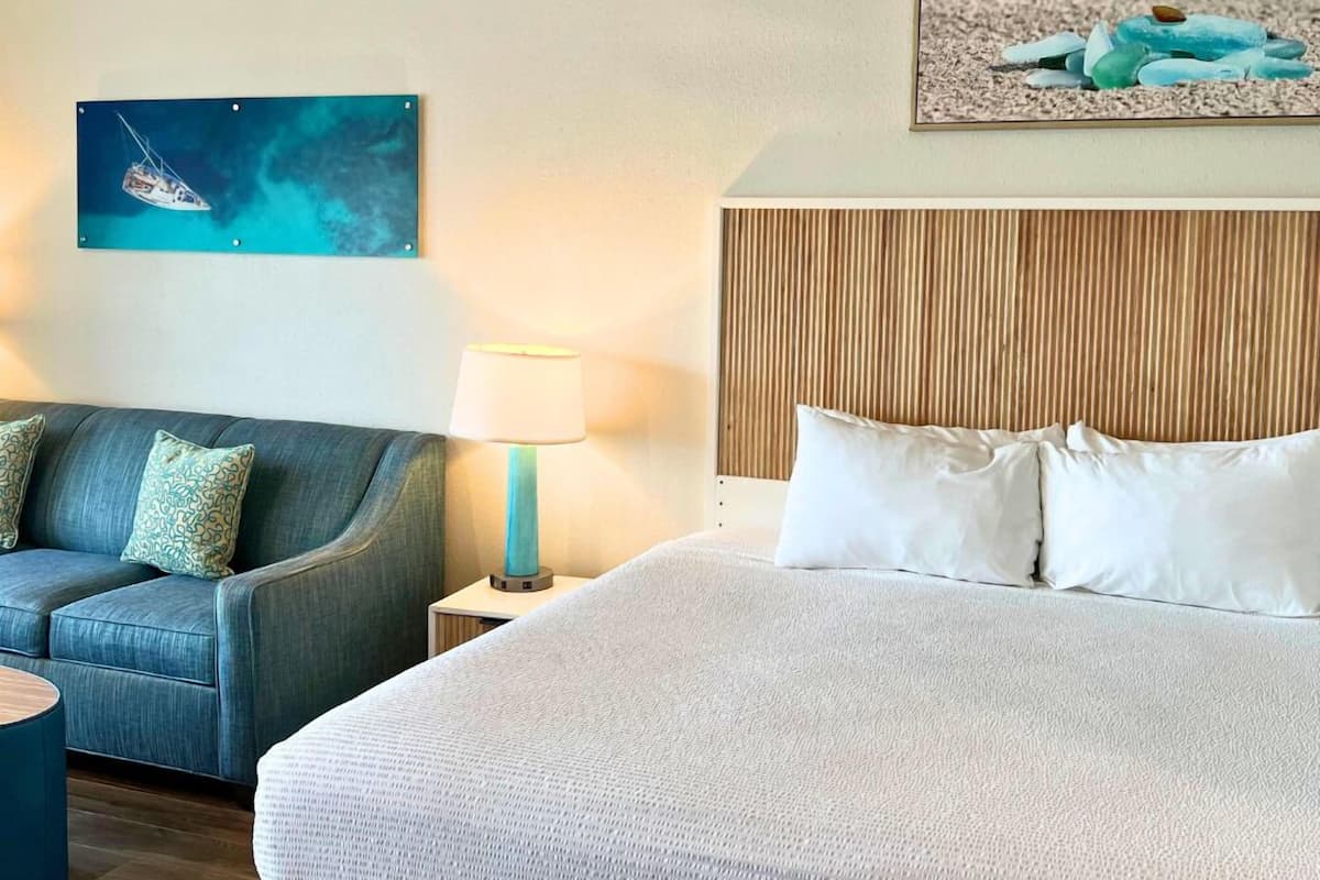 A hotel room with a large bed, a sofa, a nightstand with a lamp, and a wall with a painting of a sailboat.