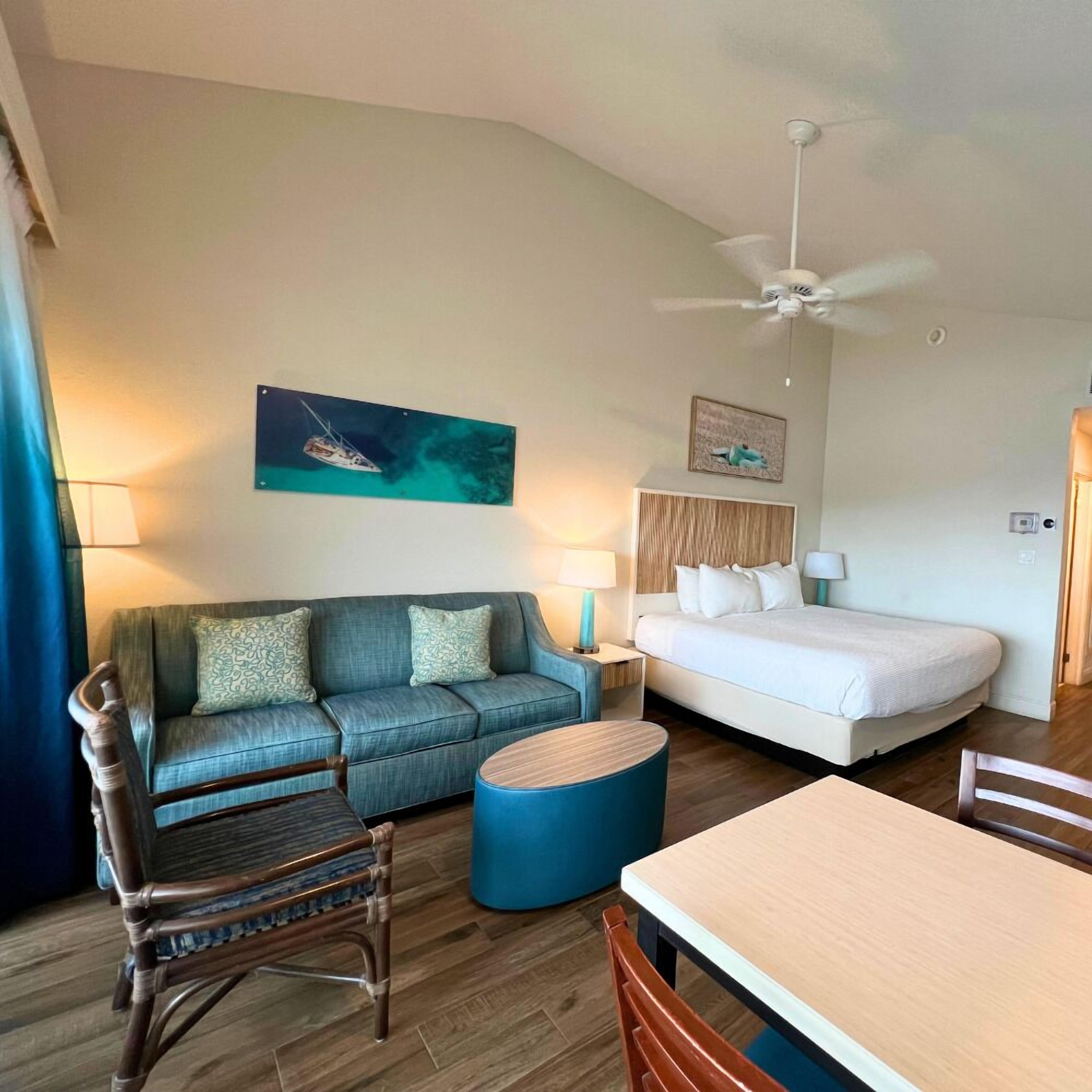A hotel room with a bed, a sofa, a dining table, a chair, a ceiling fan, and artwork on the walls.