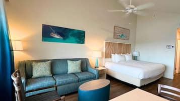 A hotel room with a bed, a sofa, a dining table, a chair, a ceiling fan, and artwork on the walls.