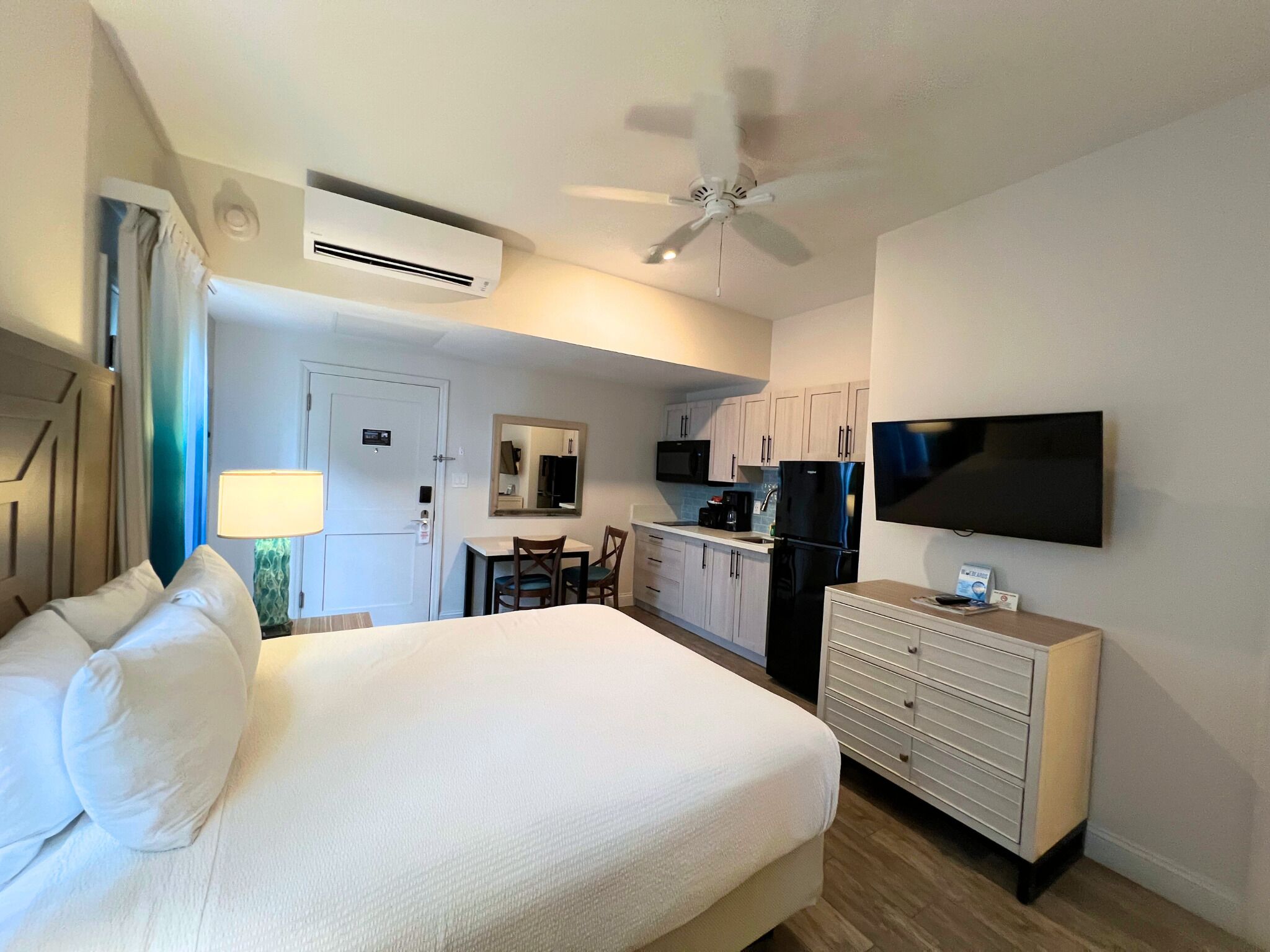 Habitación de hotel con cama, escritorio, silla, televisor, ventilador de techo y kitchenette.