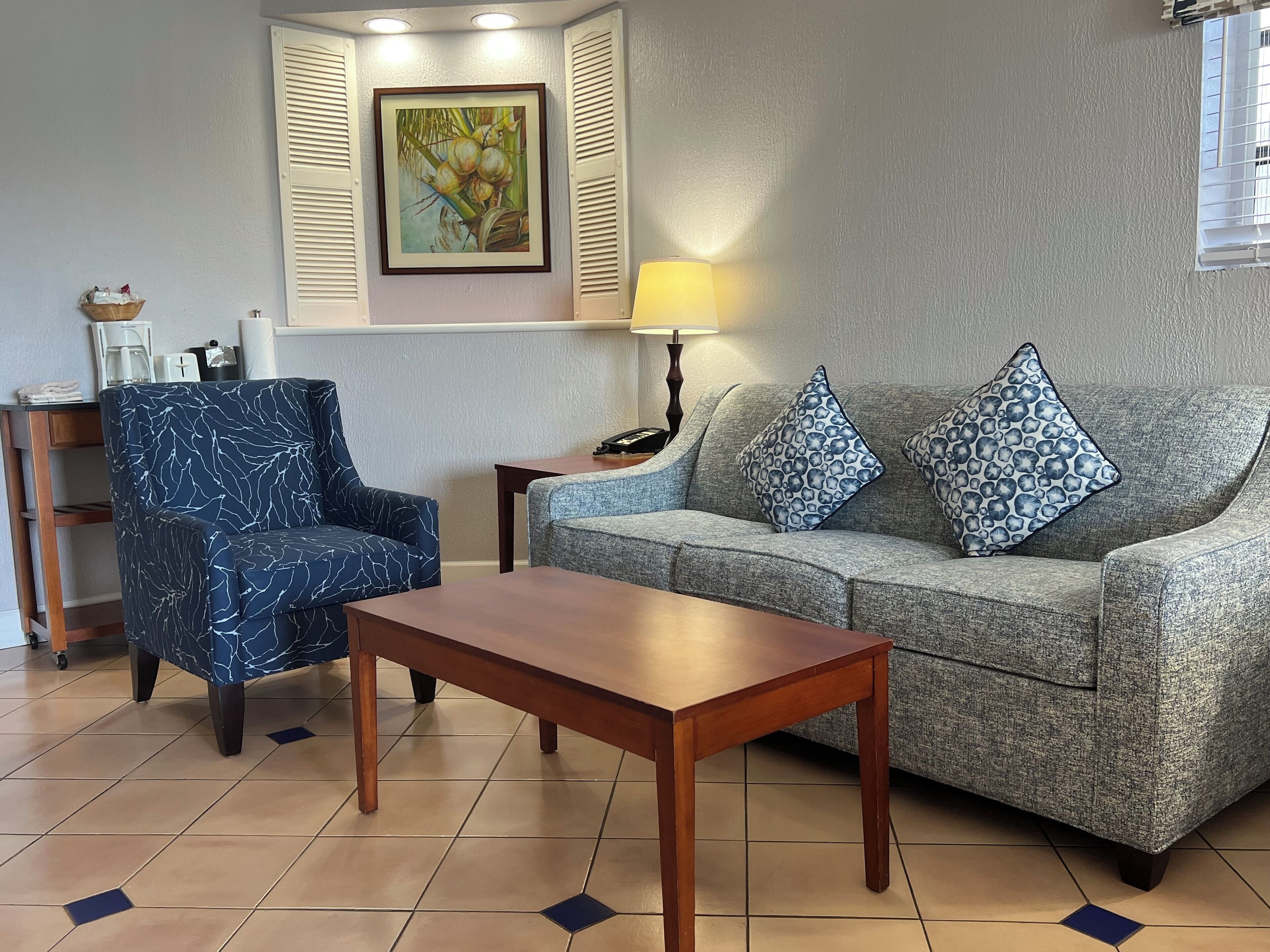 Ein Wohnzimmer mit einem blauen, gemusterten Sessel, einem grauen Sofa mit gemusterten Kissen, einem Holztisch, einer Lampe und einem Bild an der Wand.