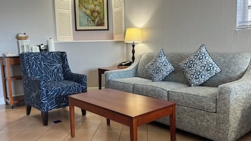 A living room with a blue patterned armchair, a grey sofa with patterned cushions, a wooden coffee table, a lamp, and a painting on the wall.