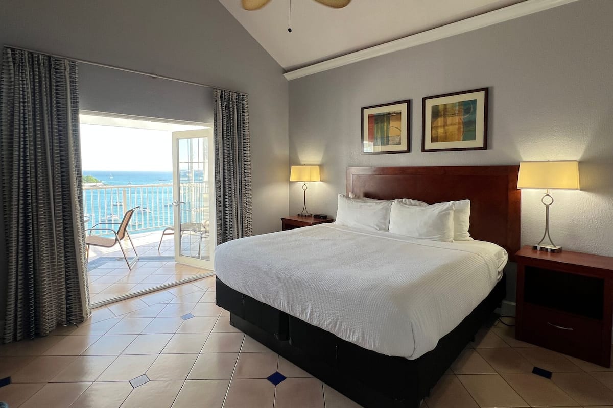 A hotel room with a large bed, two bedside lamps, a ceiling fan, and a balcony with a view of the sea.