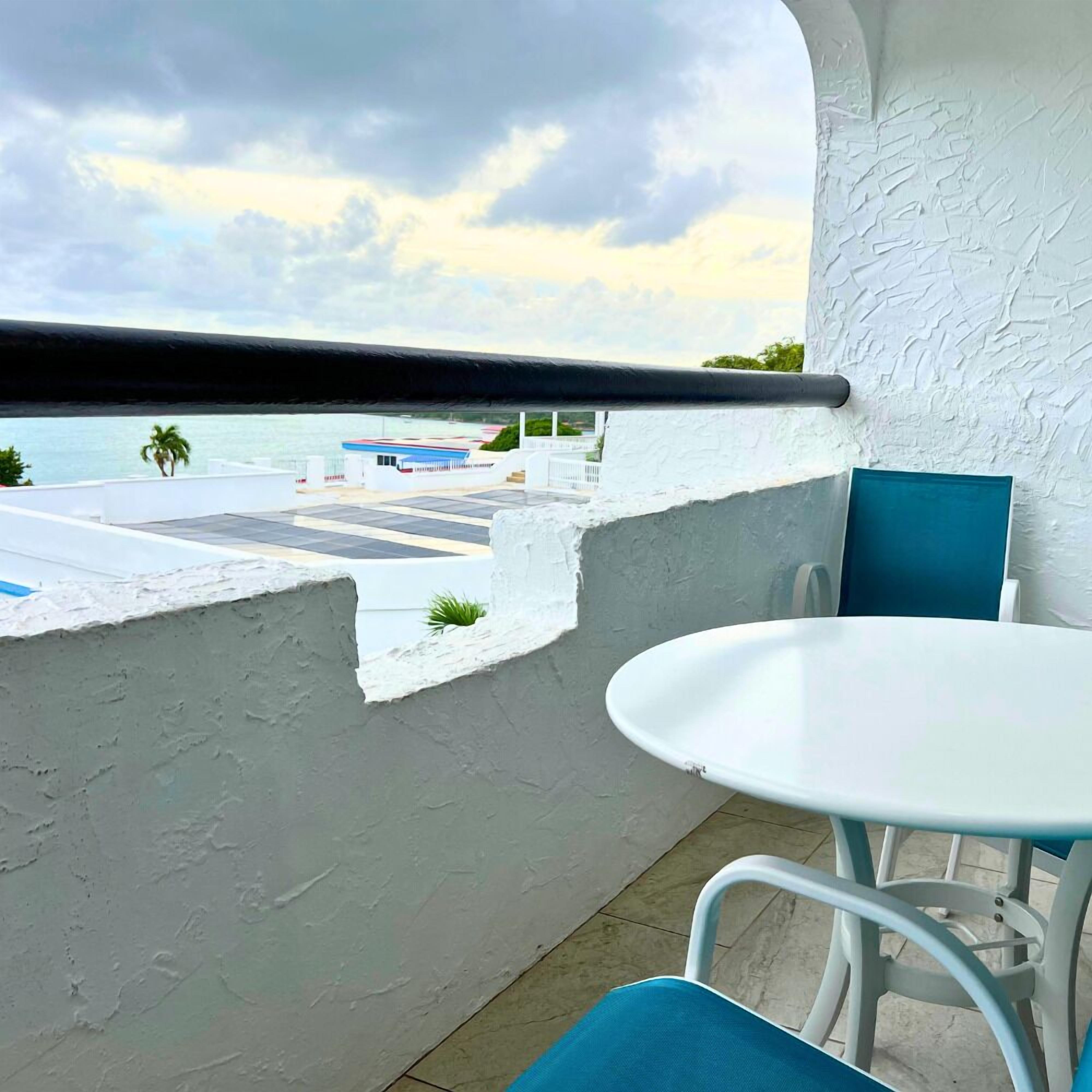 Een balkon met een witte tafel en blauwe stoelen, met uitzicht op een waterkant met gebouwen en een palmboom.