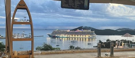 Zona de bar con cartel de cervezas, vitrina de cristalería y una vista de un crucero al fondo.