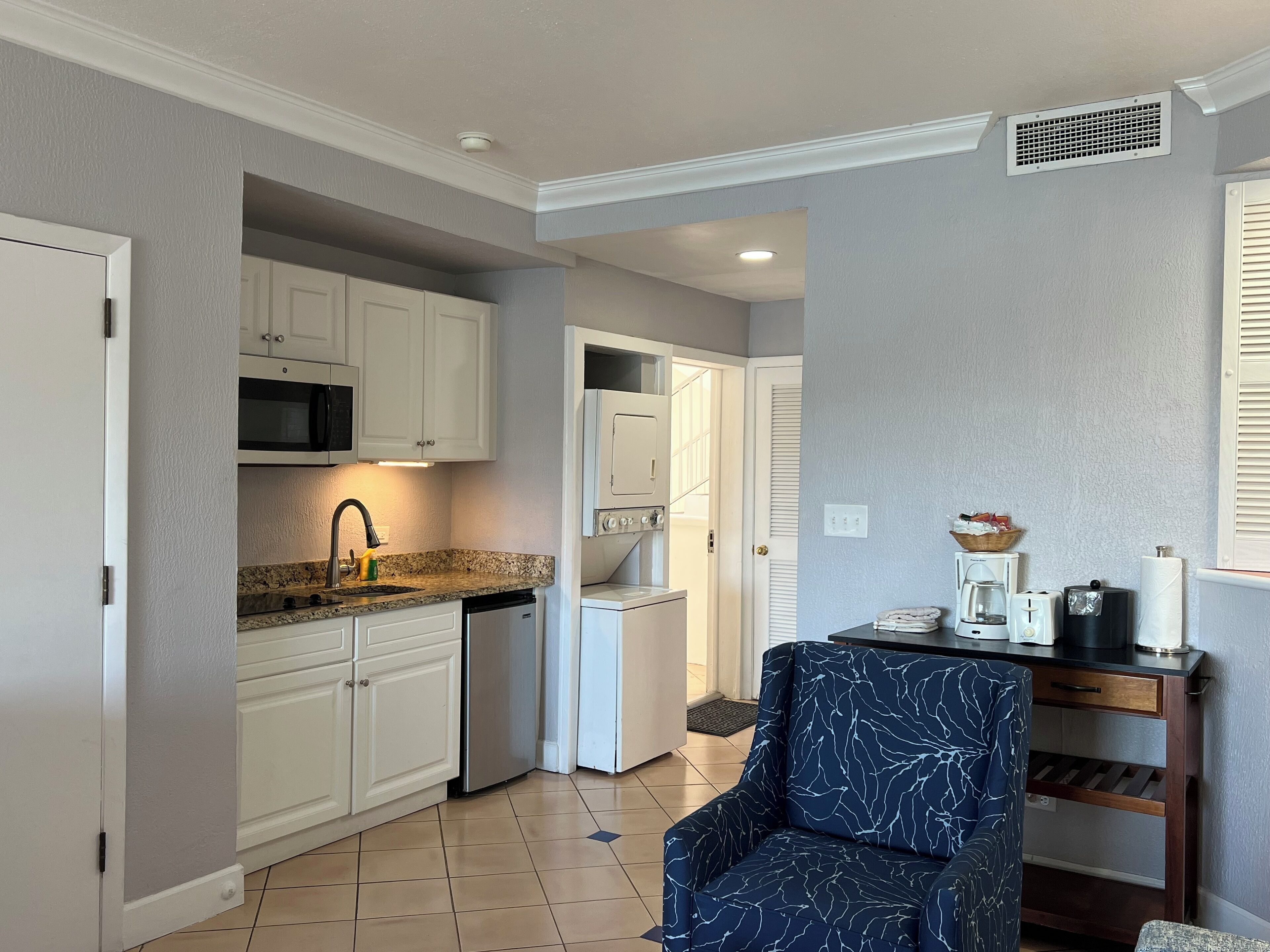 A compact kitchen with white cabinets, a microwave, and a dishwasher. There's a blue armchair and a small wooden table with a basket and a kettle.
