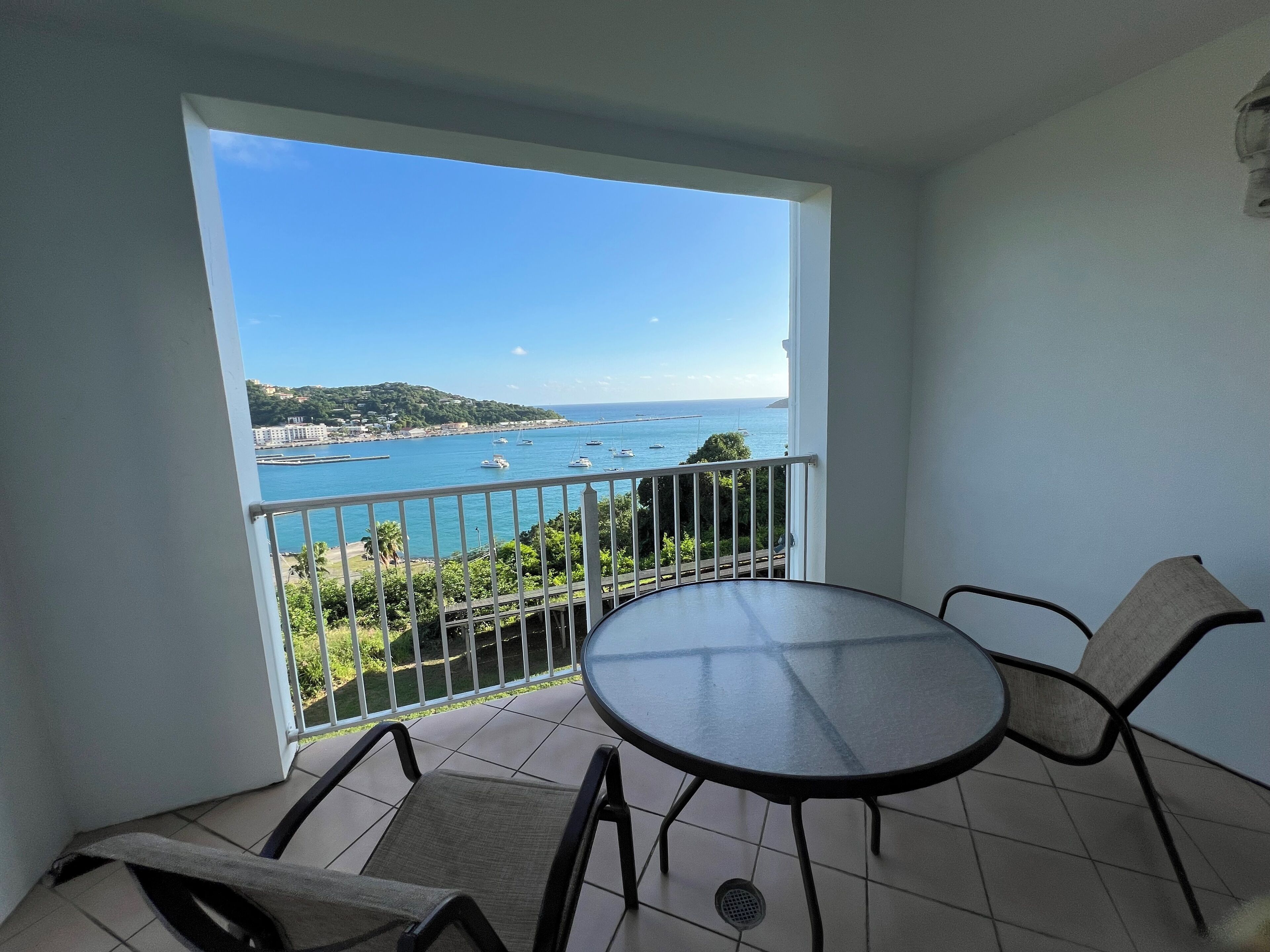 A balcony with a table and chairs, overlooking a coastal view with boats and a hilly landscape.