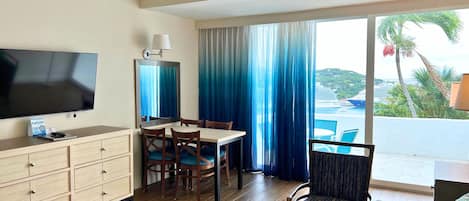 Habitación de hotel con televisión de pantalla plana, muebles de madera, una mesa de comedor y vistas a una piscina y palmeras.