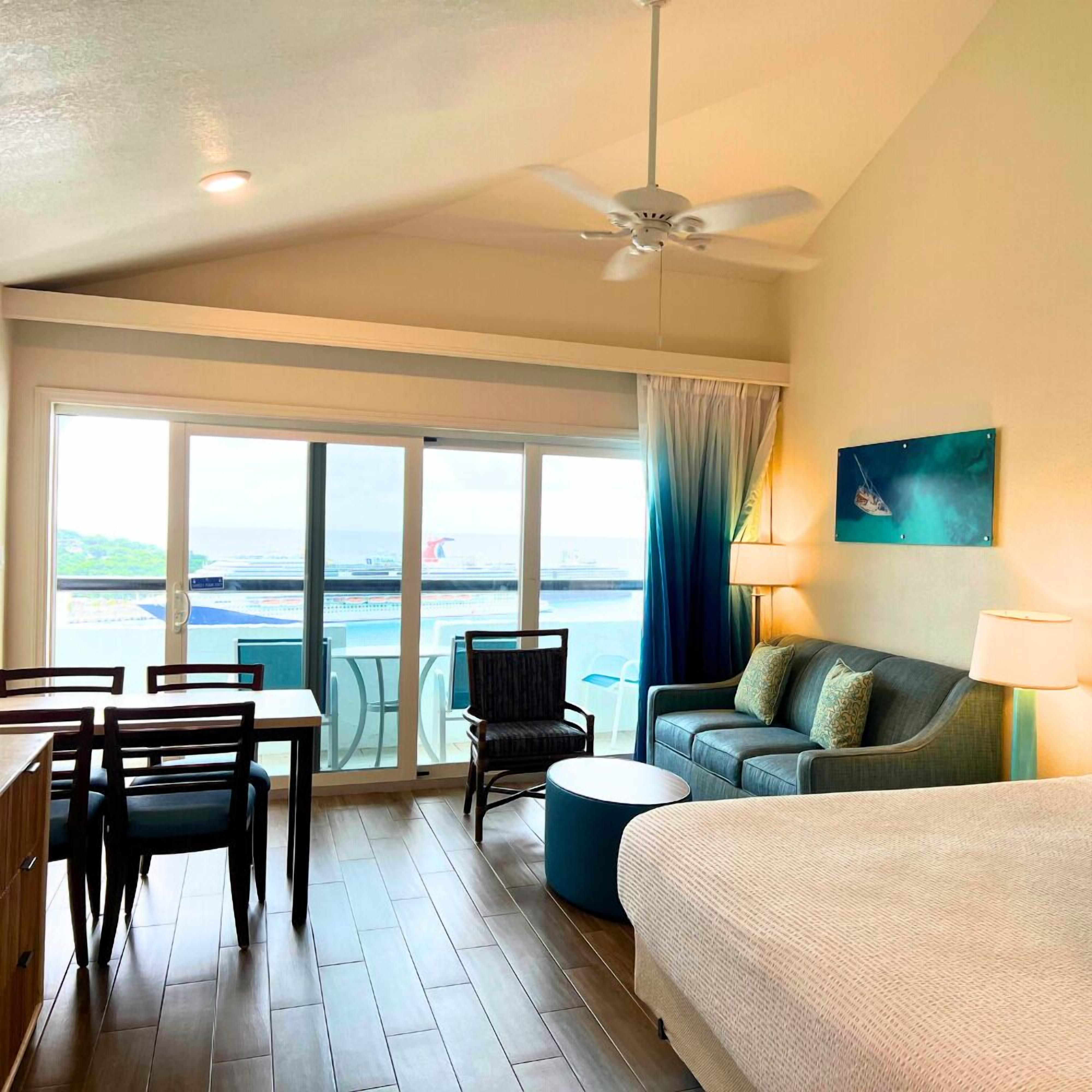 A hotel room with a balcony, a bed, a sofa, a dining table with chairs, and a ceiling fan.