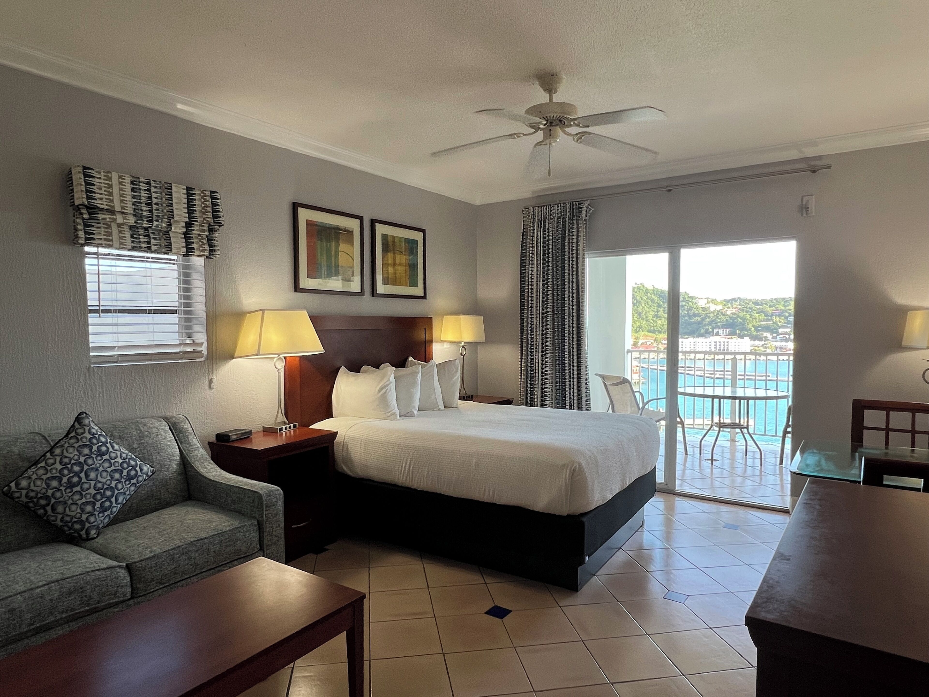 A hotel room with a bed, a sofa, a desk, a ceiling fan, a window with blinds, a door, and a view of a pool and buildings.
