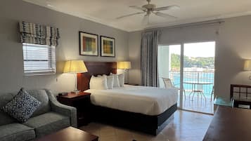 Habitación de hotel con cama, sofá, escritorio, ventilador de techo, ventana con persianas, puerta y vistas a una piscina y edificios.