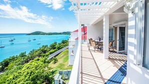 A balcony with a view of the sea, a table with chairs, and a white building with a red roof. - Bluebeard's Castle Resort (Charlotte Amalie)