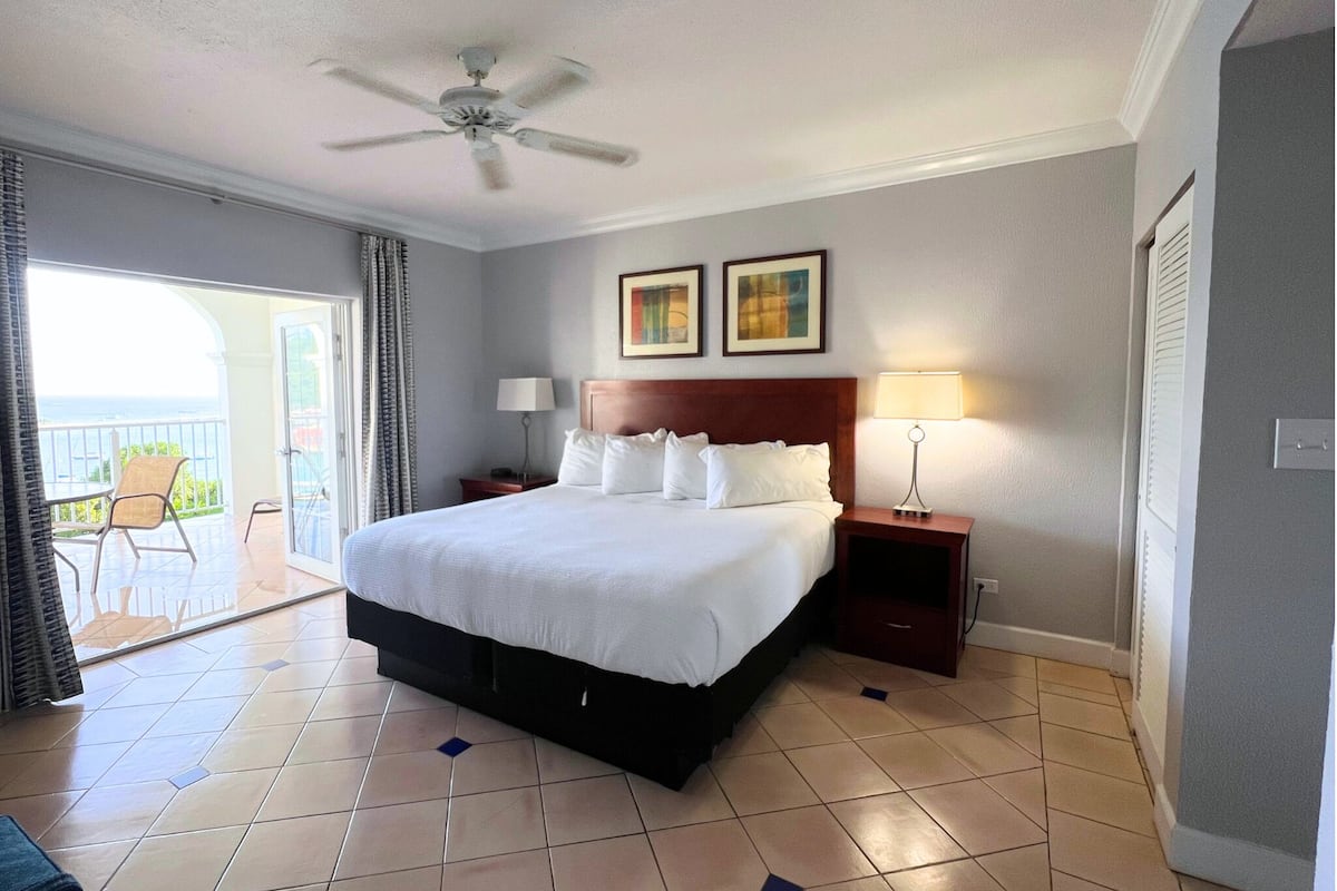 A hotel room with a large bed, a ceiling fan, two bedside lamps, a nightstand, and a view of the ocean through a sliding glass door.