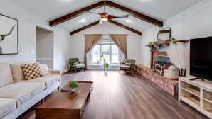 A spacious living room with a high ceiling, wooden beams, a fireplace, a television, and a large window with curtains. - Nancy`s Ranch + hot tub (Derby)
