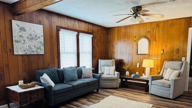 A living room with a dark blue sofa, two armchairs, a wooden ceiling fan, and a large window with blinds.