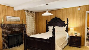 A bedroom with a dark wooden bed, a brick fireplace, a hanging light fixture, and a small bedside table with a lamp.