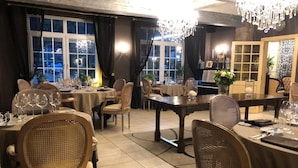Een eetruimte met meerdere tafels en stoelen, grote ramen en een kroonluchter.