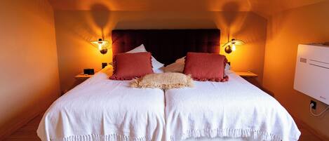 Una cama doble con ropa de cama blanca y almohadas rojas, dos lámparas de noche y un aire acondicionado de pared.