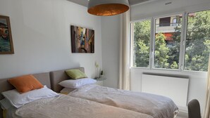 Ett modernt sovrum med två sängar, en tavla på väggen och ett fönster med utsikt över träd.