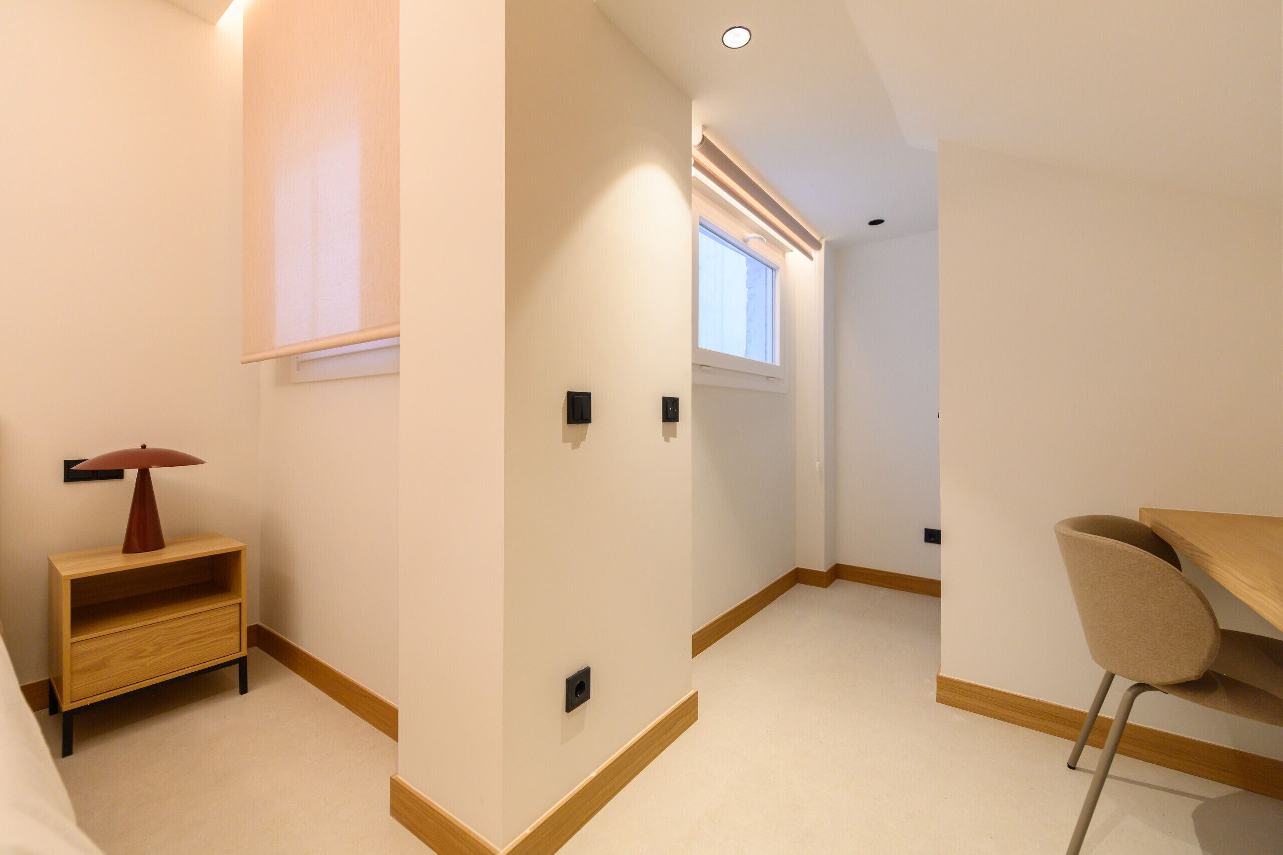 A modern, well-lit room with a wooden desk, a chair, and a small shelf with a lamp.