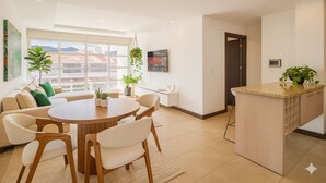 Una sala moderna con una mesa redonda de madera y sillas blancas, un sofá con cojines verdes, y una zona de cocina con encimera y plantas colgantes.