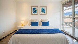Habitación de hotel con una cama grande, una mesita de noche con lámpara, dos cuadros abstractos enmarcados y un ventanal con vistas a la ciudad.