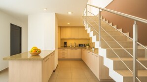 Una cocina moderna con una isla central, una escalera con barandillas de vidrio y una puerta a la izquierda.