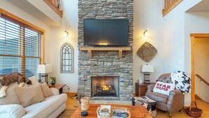 Una sala de estar acogedora con chimenea de piedra, un televisor de pantalla plana montado encima, un sofá confortable, una mesa de centro de madera con revistas y una cesta, y un perro doméstico descansando sobre el sofá.