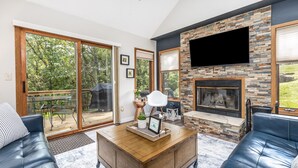 Uma sala de estar ampla com lareira, uma TV de tela plana fixada em uma parede de pedra e uma porta de vidro deslizante que dá para um pátio externo.