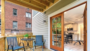 A wooden deck with outdoor seating, a sliding glass door to a kitchen, and a brick building in the background.