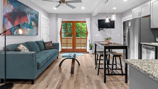 A modern kitchen with a built-in oven, a dining area with a wooden table and bar stools, a ceiling fan, and a large window with a view of greenery.
