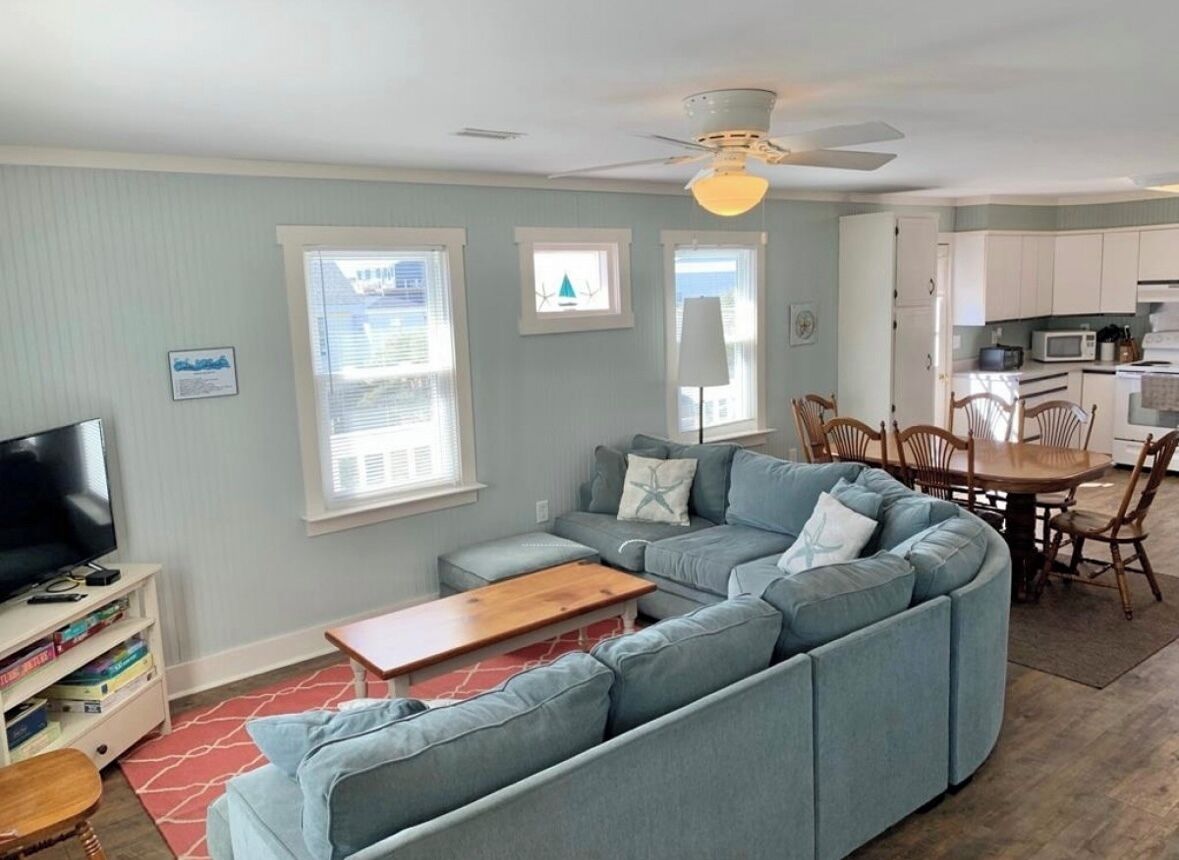 A living room with a sectional sofa, a wooden coffee table, and a TV stand. There is a dining area with a table and chairs.