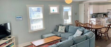 A living room with a sectional sofa, a wooden coffee table, and a TV stand. There is a dining area with a table and chairs.