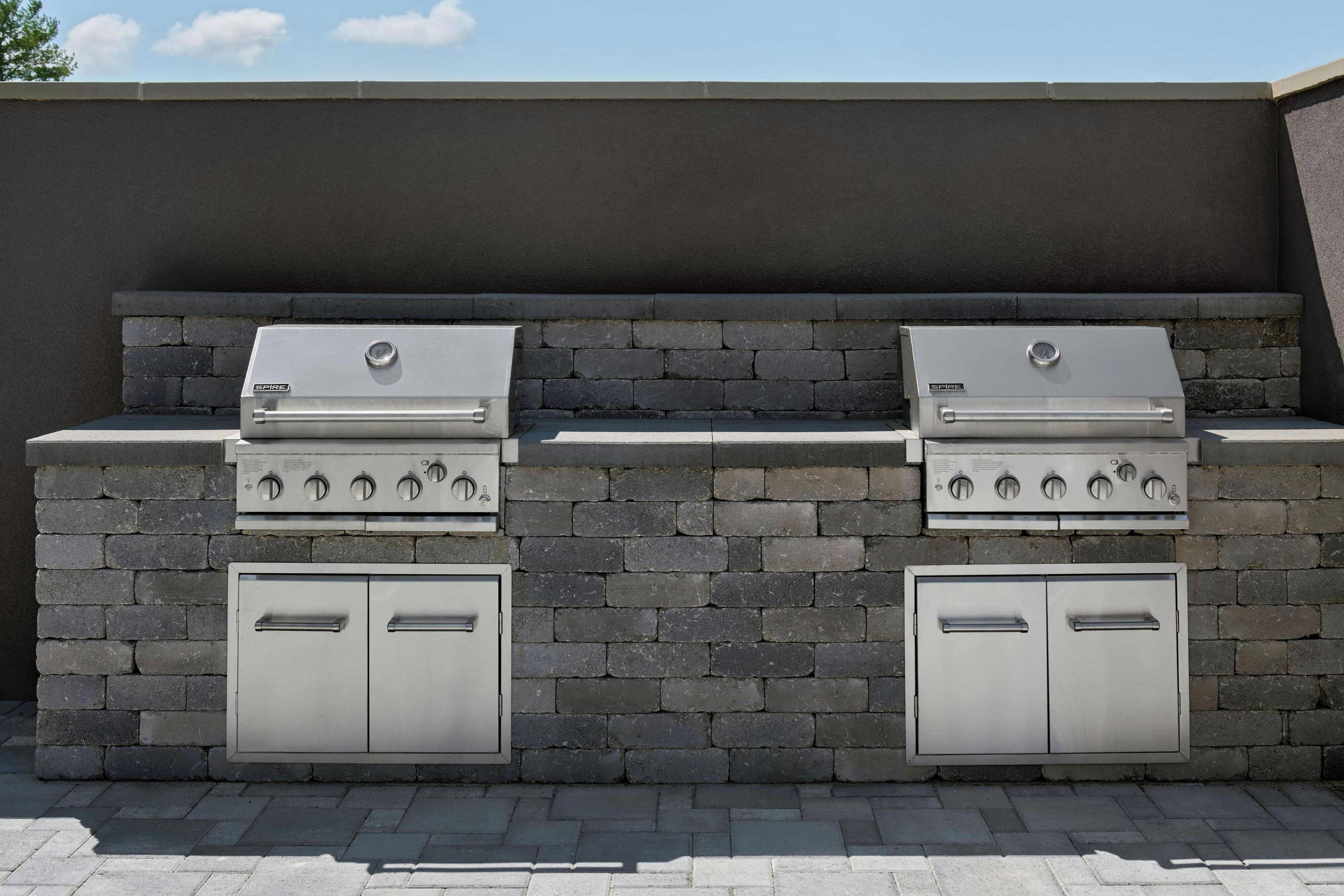 A barbecue grill setup with two gas grills and two built-in cabinets against a stone wall.