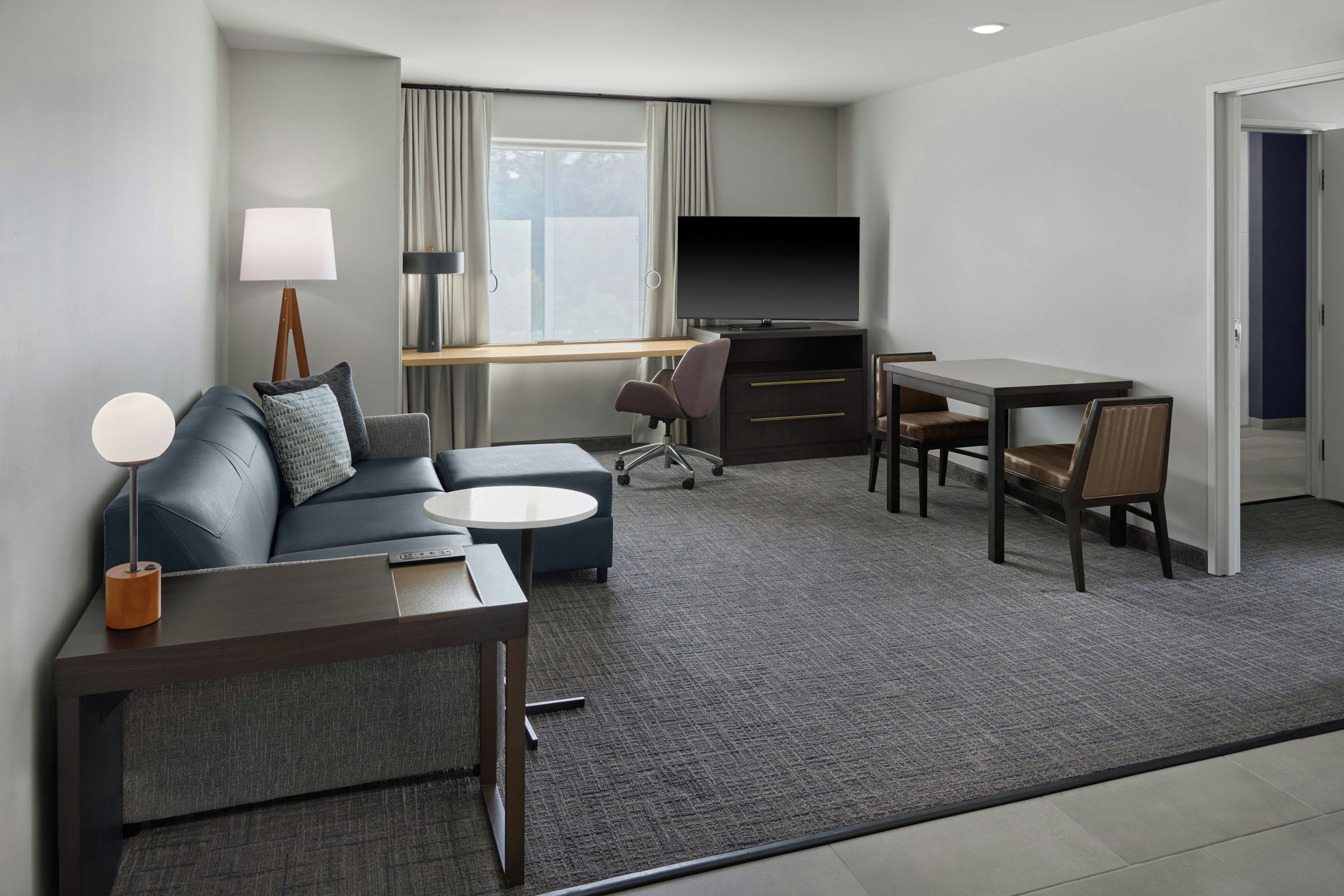 A modern hotel room with a sofa, a small round table, a desk with a chair, a television, and a lamp.