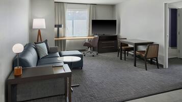 A modern hotel room with a sofa, a small round table, a desk with a chair, a television, and a lamp.