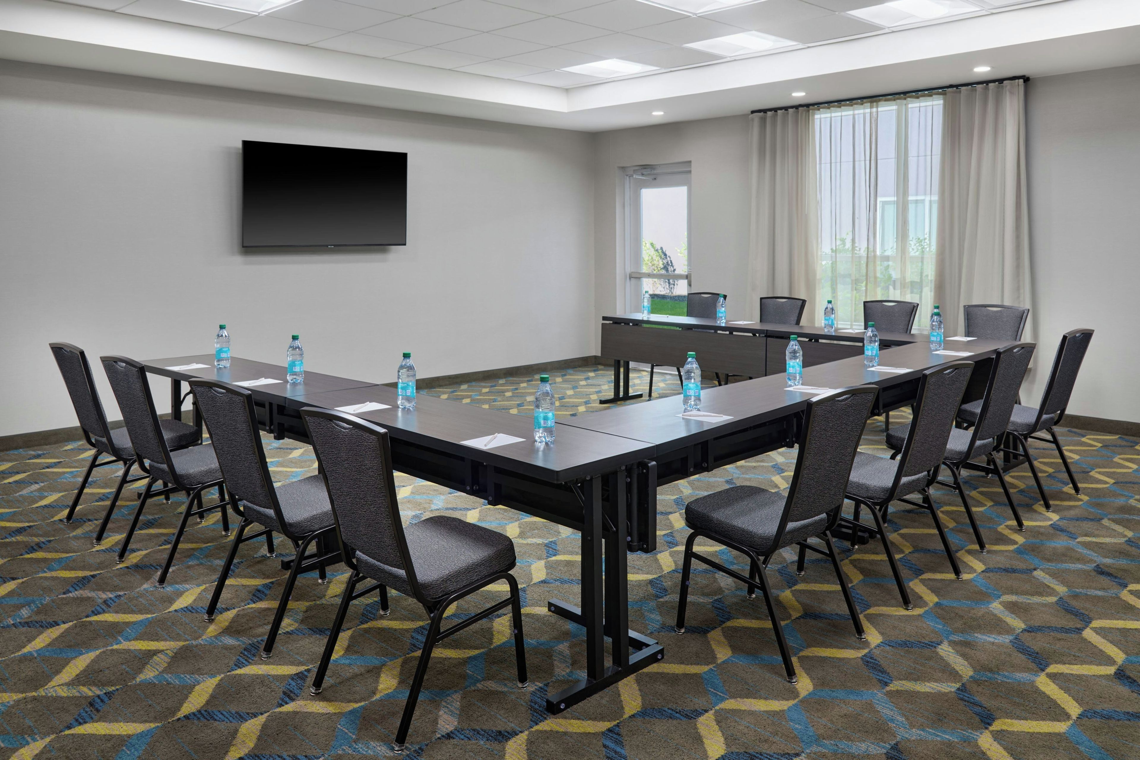 Un salón de conferencias con una larga mesa, sillas, botellas de agua, un televisor de pantalla plana y una ventana con cortinas.