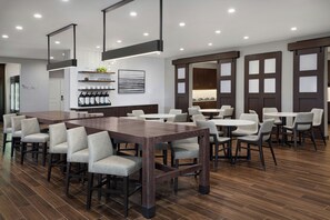 A modern dining area with a long wooden table and multiple chairs, a coffee bar with various coffee-making equipment, and a kitchen area with cabinets and a refrigerator. - Residence Inn By Marriott Traverse City (Traverse City)