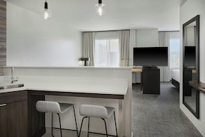 A modern hotel room with a kitchen area, bar stools, a flat-screen TV, and a large window offering a city view.