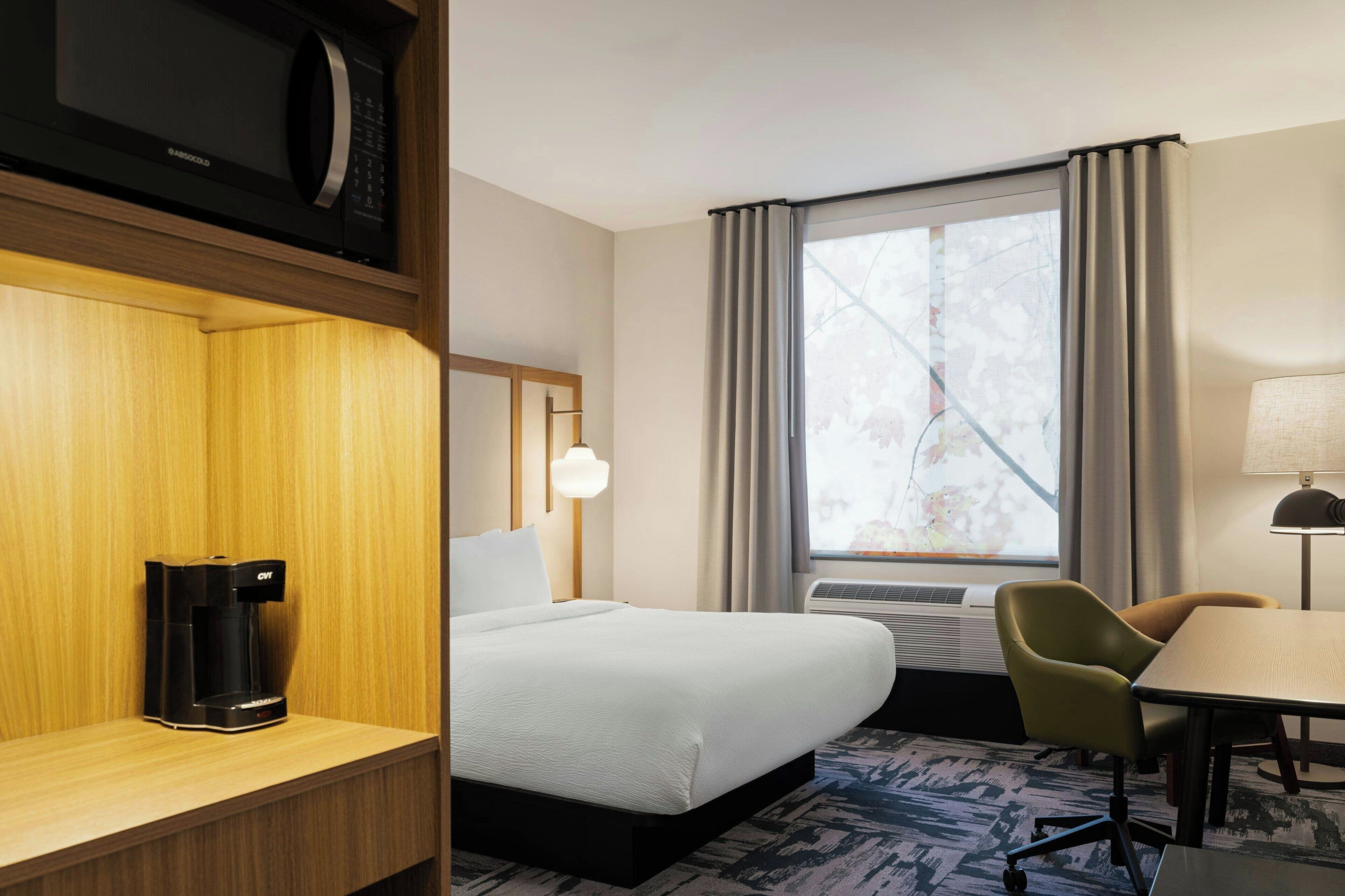 A hotel room with a bed, a desk with a coffee maker, a chair, a lamp, and a window with curtains.