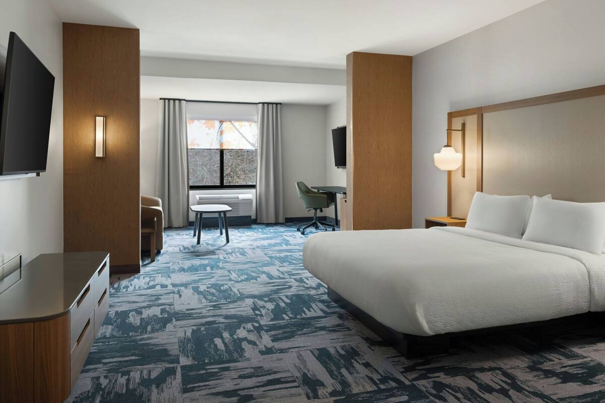 Quarto de hotel com uma cama grande, uma escrivaninha, uma cadeira, uma televisão e uma janela com cortinas.