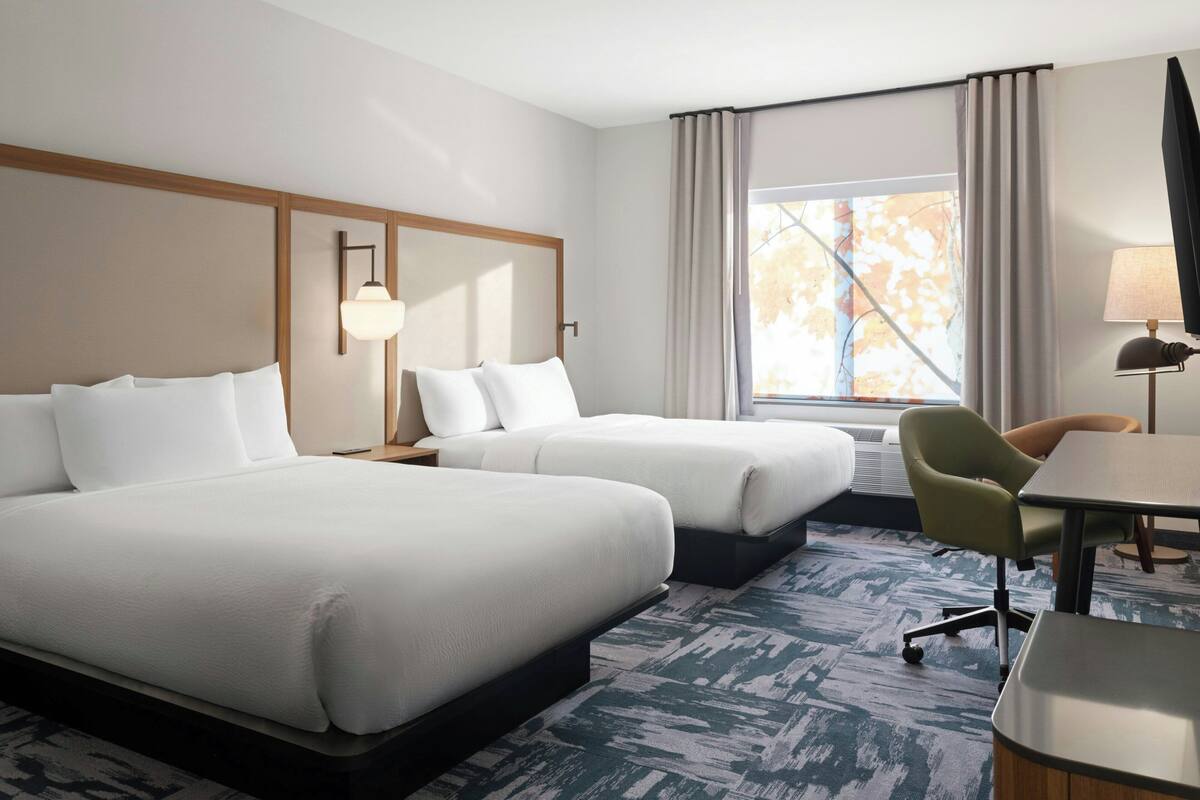 Quarto de hotel com duas camas, uma escrivaninha, uma cadeira, um abajur e uma janela com cortinas.