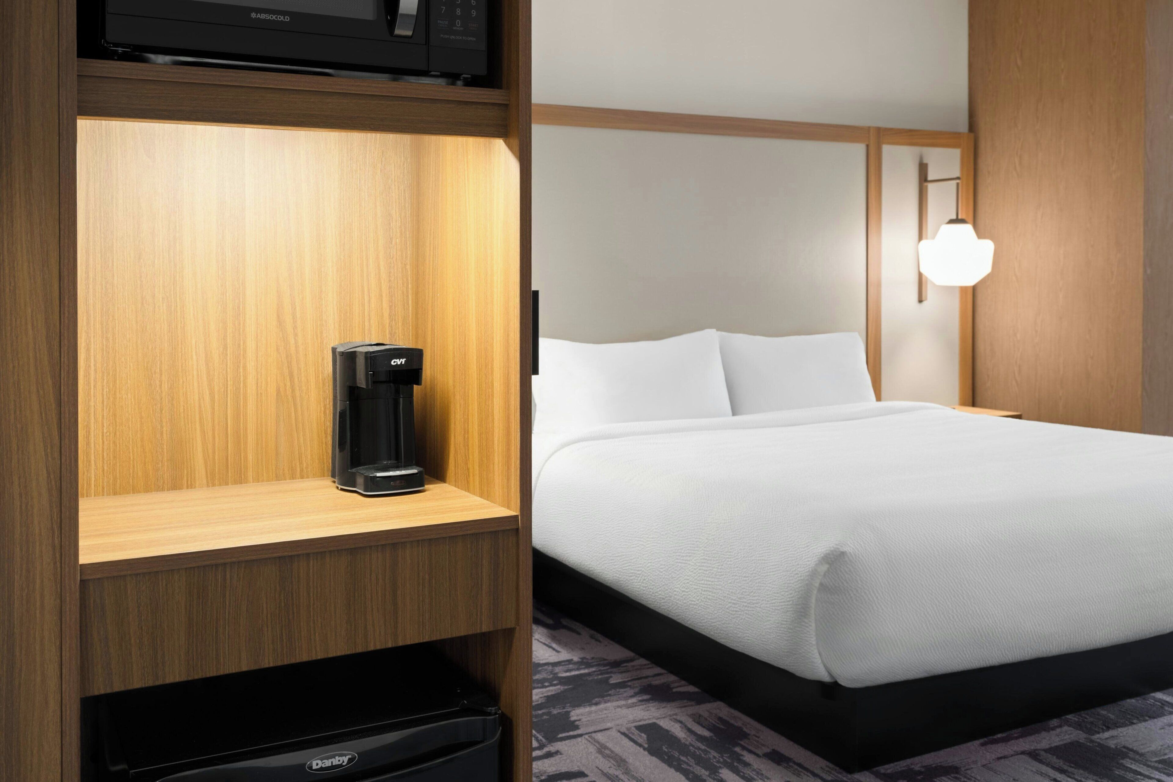 A hotel room with a bed, a coffee maker on a wooden shelf, a black trash bin, and a wall-mounted light fixture.