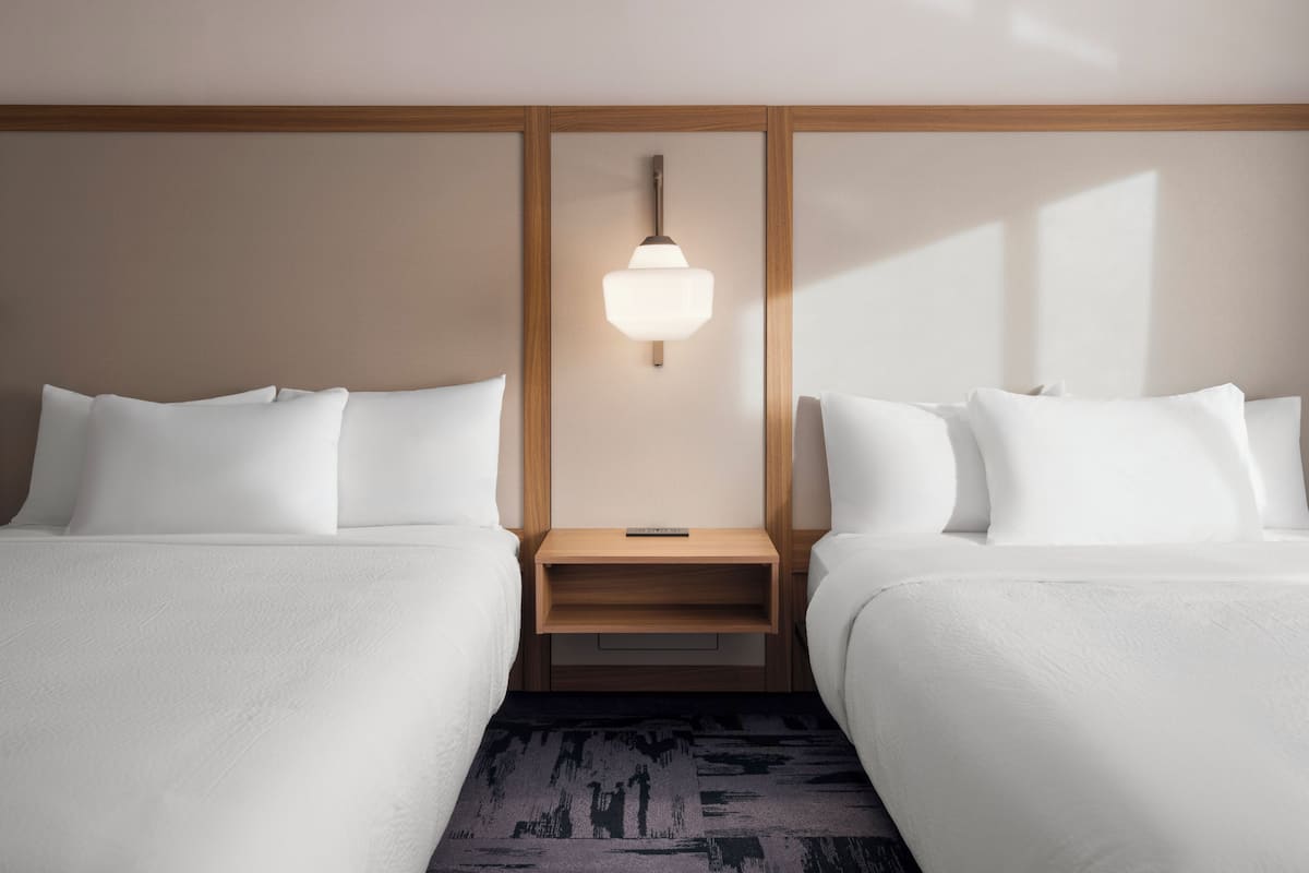 Quarto de hotel com duas camas, uma mesa de cabeceira, um abajur e um tapete estampado.