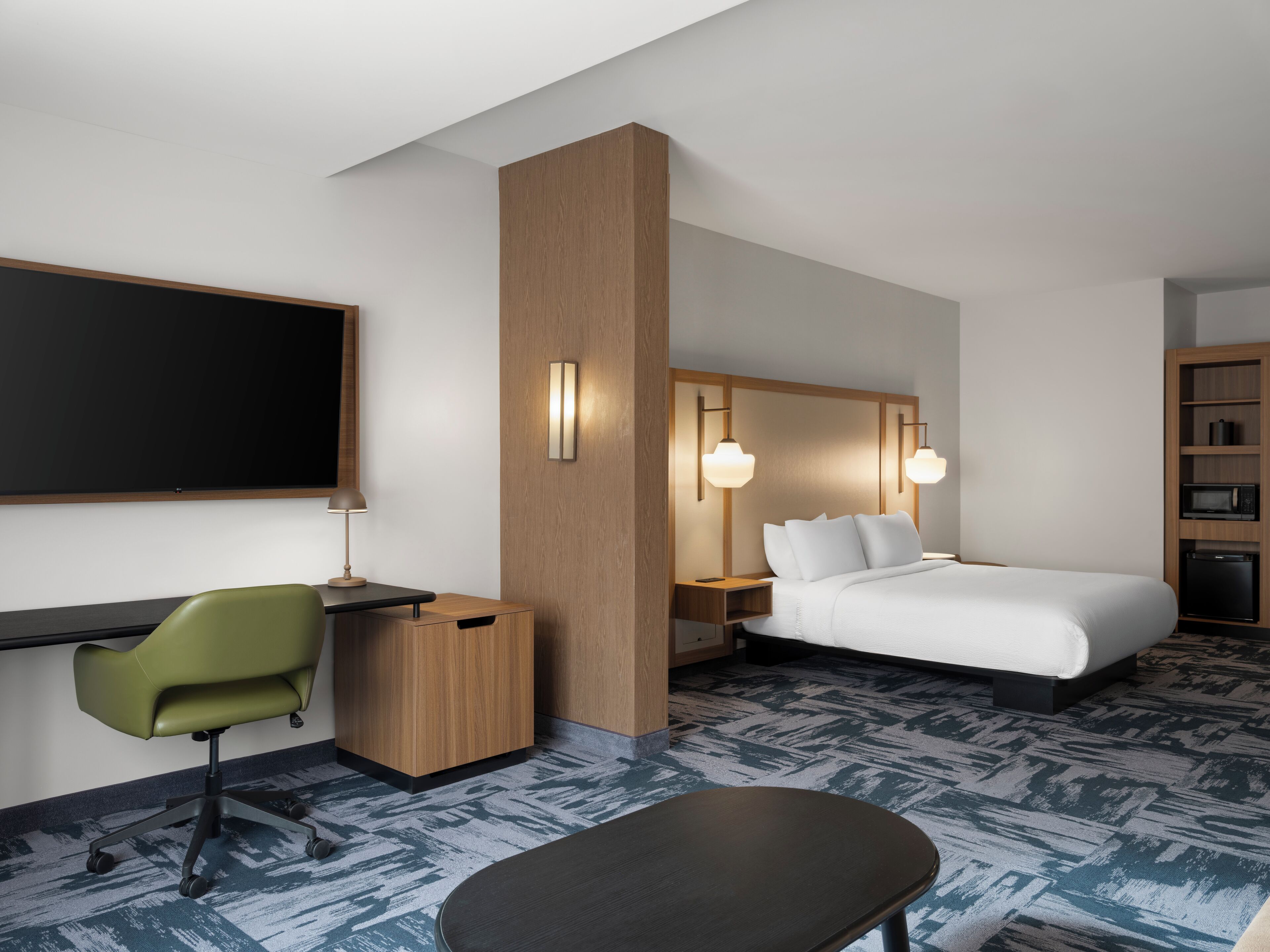 Habitación de hotel con escritorio, una silla verde, cama, mesitas de noche y un microondas.